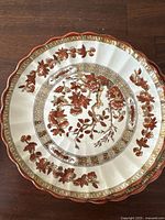 Close-up of porcelain saucer with intricate orange/red floral India Tree pattern and scalloped edges with gold trim