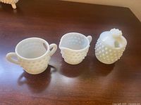 Front view of three white hobnail milk glass items: sugar bowl with two handles, creamer with handle, honey bowl with lid.