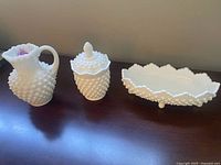 Three white hobnail milk glass pieces arranged side by side: mini pitcher, covered candy dish with lid, oblong candy dish.