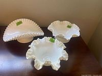 Photo of three white hobnail milk glass pieces on a dark wood surface: an open basket with ruffled edge and two smaller wavy edge bowls (round and scalloped)