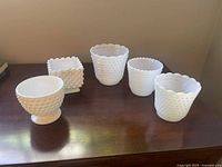 Five assorted hobnail milk glass vases on dark wooden table, showing variety of shapes including a square and a footed vase.