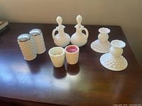 Overview of all hobnail milk glass items: salt and pepper shakers, cruets, small cups, and candle holders arranged on a wood surface.
