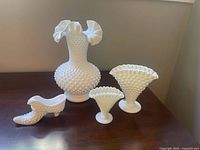 Front-angle photo of all four hobnail milk glass pieces on a wooden surface including the large vase, shoe figurine, and two wavy triangle vases.