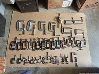 Lot of 37 small metal clamps, laid out on cardboard, showing varying sizes of C-clamps and a few bar clamps with metal screw tightening mechanisms.