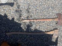 Photo showing two vintage axes laid on gravel, one with a long wooden handle and single blade, the other shorter with a double blade.