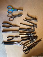 Top-down view of 11 vintage hand tools arranged on cardboard on the floor. Tools include scissors, pliers, pipe wrench, punch tool, and tin snips.