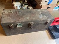 Closed view of a vintage metal toolbox with rusted and worn lid showing faded stencil markings and two metal latch locks, one with a lock plate.