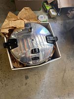 Top-down view of the large round shiny metal pressure cooker lid featuring black side handles and a pressure valve on top, placed inside a cardboard box with brown paper packing.