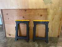 Two folded black and yellow Craftsman sawhorses leaning against plywood wall inside a garage.