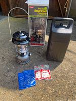 Full lot view showing Coleman lantern, carrying case, original box, and replacement mantles on the ground in sunlight.