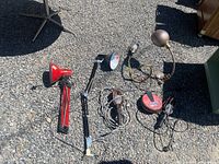Six assorted vintage metal shop lamps laid out on gravel, showing a variety of clamp, desk, and adjustable arm styles mostly in red or brown metal finishes with cords attached.