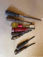 Photo shows a collection of vintage screwdrivers and hand tools on a cardboard surface, featuring a variety of wooden and plastic handles with visible wear and paint marks.
