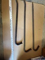Three vintage steel crow bars laid out on cardboard next to a ruler showing varying lengths around 18 inches.