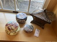 Four decorative boxes arranged on a surface near window light: round ceramic box, round glass box, small round resin box, and large ornate resin box with pyramid lid