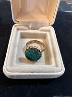 Ring displayed in a cream-colored jewelry box, top-down view showing bezel-set green stone and gold band.