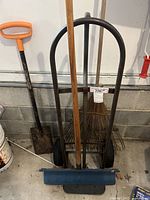 Hand truck, rake, pitch fork, and shovel leaning against wall