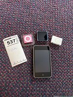 Four Apple iPods arranged on carpet including a black front iPod Classic, a pink iPod Shuffle in case, blue iPod Nano, and white iPod Shuffle in case.