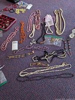 Overview of multiple necklaces, some with faux pearls in colors including white and lavender, polished stone beads in orange and black, rhinestone earrings, and small jewelry pieces.