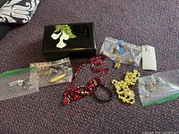 Overview of the lot items including multiple beaded necklaces, small bags with pins and earrings, and decorative black box with white flowers on top lying on carpeted floor.