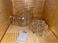 Pitcher and bowl placed on wooden shelf with identification tag. Pitcher is round and clear with spout and handle; bowl is smaller and geometric with visible crack.