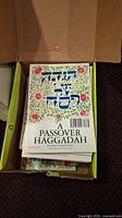 Box of sixteen Passover Haggadah books with decorated front covers visible in the box.