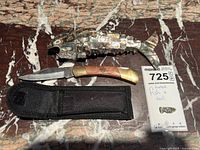 Photo showing the jointed abalone shell fish sculpture alongside the folding Boy Scout cutter knife and its black nylon sheath on a marble surface, with a note indicating measurements.