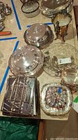 Photo of assorted silver plated covered dishes, pitcher, glass bowl with metallic rim, and a toast rack on a table.