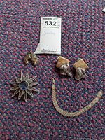 Five pieces of sterling jewelry arranged on multicolor carpet: mesh bracelet, two pairs of earrings (clip-on and pierced), and a flower-shaped brooch with a blue center stone.
