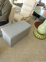 Side angle view of one light gray upholstered storage bench placed on beige carpet, showing cushioned top and fabric texture.