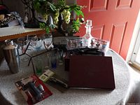 Photo showing the full barware set including hand-painted wine glasses, shot glasses in various colors, a stainless steel cocktail shaker, a metal bottle holder, cork stoppers in a display, a wooden box, and cocktail tools.