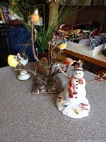 Metal tree with removable bird figurines in multiple colors and Lenox snowman figurine with plaid scarf and black top hat decorated with floral accents.