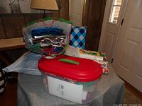 Photo showing a large plastic storage container with a red lid filled with gift wrapping materials. Multiple gift bags and rolls of wrapping paper are placed on and around the container.