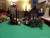 Wide shot showing two decorative black metal lanterns on green tablecloth, decorated with artificial pine branches, pinecones, red berries, and spherical Christmas ornaments.