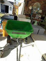 Front view of the green John Deere spreader showing the plastic hopper, metal frame, and wheels, outdoor setting with furniture in background.
