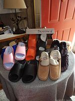View of front row shoes including pink, black, orange slides, beige moccasins, and patterned slippers along with Bearpaw box on top.