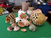 Photo showing two ceramic fish-shaped votive candle holders, various decorative seashells including a white conch shell and a flower decoration, all arranged on a green surface.