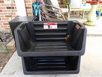 Two black Husky stackable storage totes displayed one on top of the other outside on a patio floor. The totes are new and made of reinforced plastic with ribbed bottoms.