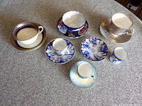 Top-down view of seven assorted porcelain teacups and saucers in blue, white, purple and yellow tones on gray woven fabric surface.