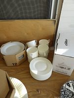 Photo showing white plates, ceramic mugs, and saucers on a wooden surface with boxes.
