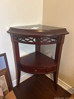 Front angled view of the cherry wood tone curved corner accent table showing its curved top, open fretwork trim, lower shelf, and drawer.