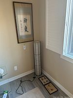 The cylinder floor lamp positioned in a corner next to a window, with one large framed Asian character print hanging on the wall above and two smaller prints on the floor below the lamp.