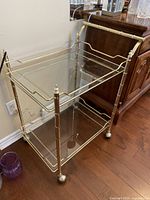 Front three-quarter view of bar cart showing brass finish frame, upper and lower glass shelves, caster wheels.