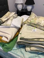 Photo showing folded comforter, pillows, blankets in multiple colors on mattress with headboard and lamps in background