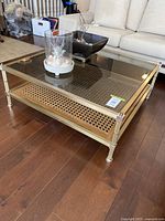 Three-quarter view of square coffee table showing brass metal frame, smoked glass top and wood-cane lower shelf in living room setting