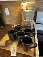 Proctor Silex coffee maker with 5 dark grey mugs arranged on a glass cutting board with fruit design