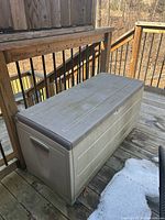 Plastic Keter deck box sitting outdoors on a wooden deck, showing one long side of box with beige body and taupe lid, with visible dirt and staining from outdoor use.