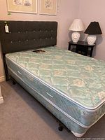 Full view of bed with mattress, box spring and grey buttoned fabric headboard against wall with two night lamps.
