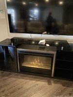 Full view of the black glass electric fireplace with silver trim installed under a mounted TV. Fireplace shows flame effect active. Glass door appears slightly misaligned or detached on one side.