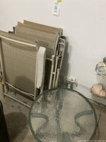Four stacked beige metal framed folding chairs with fabric seats and backs, showing armrests and foldable frames, photographed leaning against a wall indoors.