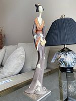 Full view of the slender 17 inch tall geisha resin figurine positioned on a glass table next to a lamp and sofa.
